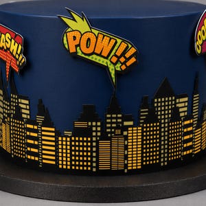 Puede incluir: Un pastel azul oscuro decorado con una silueta de ciudad negra con ventanas amarillas. En la parte superior hay globos de texto estilo c&oacute;mic con las palabras CRASH, POW y OOOPS en una tipograf&iacute;a gr&aacute;fica, llamativa y colorida.