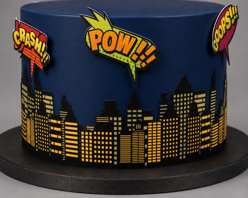 Puede incluir: Un pastel azul oscuro decorado con una silueta de ciudad negra con ventanas amarillas. En la parte superior hay globos de texto estilo c&oacute;mic con las palabras CRASH, POW y OOOPS en una tipograf&iacute;a gr&aacute;fica, llamativa y colorida.