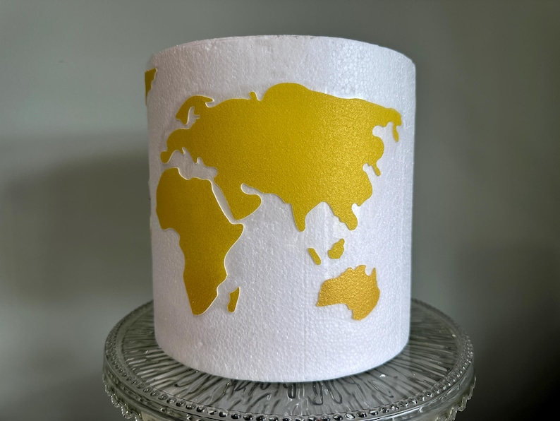 May include: A white cylinder with a gold world map design. The cylinder is on a clear glass base.