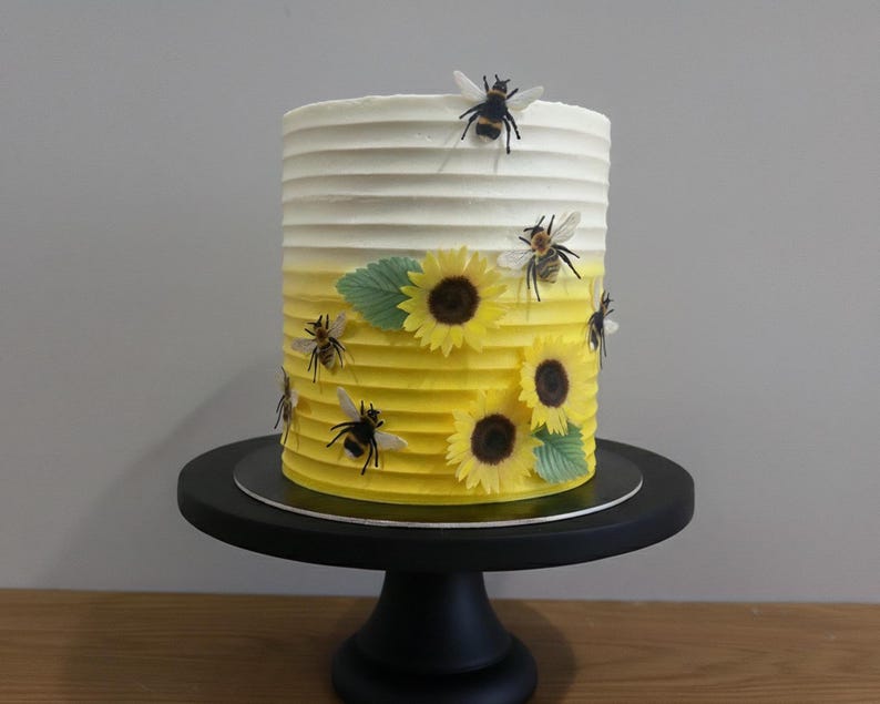 Pu&ograve; includere: Una torta cilindrica con una consistenza a righe orizzontali, caratterizzata da una glassa sfumata dal bianco al giallo. La torta &egrave; decorata con diverse piccole figure di api e tre girasoli gialli con centri marrone scuro e foglie verdi.