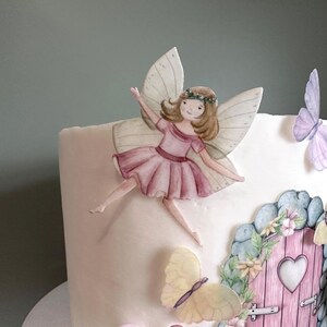 May include: A white cake decorated with a fairy figurine, butterflies, and a pink door with a heart-shaped window. The fairy has pink wings, a pink dress, and brown hair. The butterflies are yellow, purple, and blue.