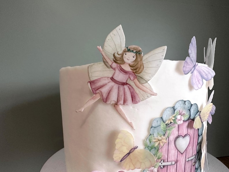 May include: A white cake decorated with a fairy figurine, butterflies, and a pink door with a heart-shaped window. The fairy has pink wings, a pink dress, and brown hair. The butterflies are yellow, purple, and blue.