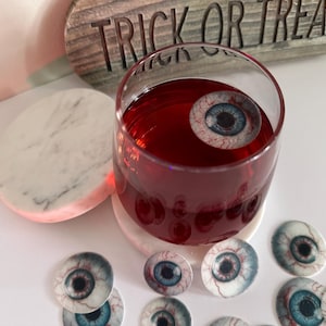 Puede incluir: Un vaso de l&iacute;quido rojo con un ojo falso flotando en &eacute;l. El vaso est&aacute; sobre un posavasos de m&aacute;rmol blanco. Hay varios ojos falsos m&aacute;s esparcidos alrededor del vaso.