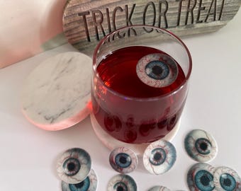 May include: A glass of red liquid with a fake eyeball floating in it.  The glass is on a white marble coaster.  There are several more fake eyeballs scattered around the glass.