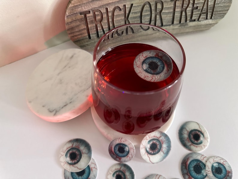 Puede incluir: Un vaso de l&iacute;quido rojo con un ojo falso flotando en &eacute;l. El vaso est&aacute; sobre un posavasos de m&aacute;rmol blanco. Hay varios ojos falsos m&aacute;s esparcidos alrededor del vaso.