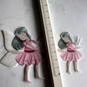 K&ouml;nnte beinhalten: Zwei Papierfeen-Ausschnitte mit rosa Kleidern, Fl&uuml;geln und Blumenkr&auml;nzen. Ein Lineal mit Zentimetermarkierungen wird neben den Feen platziert. Die Feen sind etwa 7,6 cm gro&szlig;.