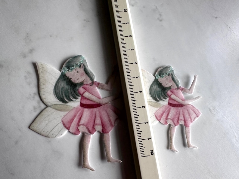 K&ouml;nnte beinhalten: Zwei Papierfeen-Ausschnitte mit rosa Kleidern, Fl&uuml;geln und Blumenkr&auml;nzen. Ein Lineal mit Zentimetermarkierungen wird neben den Feen platziert. Die Feen sind etwa 7,6 cm gro&szlig;.