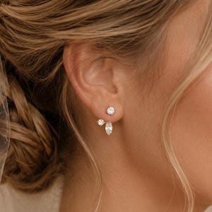 Peut inclure: Une boucle d'oreille de type ear jacket argentée composée d'une petite pierre ronde transparente sur le dessus et d'une pierre transparente taille marquise en dessous, placée derrière le lobe. Portée avec un voile blanc orné de petites perles.