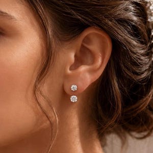 May include: A pair of double-sided stud earrings featuring two round, clear, brilliant-cut stones. The smaller stone sits on the earlobe, while the larger stone is positioned just below the lobe, creating a layered, suspended effect on the ear.