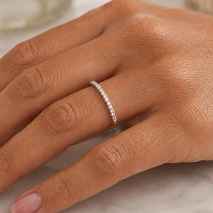 May include: A thin silver band ring featuring a row of small, clear, round-cut stones set along the top half of the band. The metal has a polished, reflective finish, and the stones are arranged in a continuous line, creating a delicate appearance.
