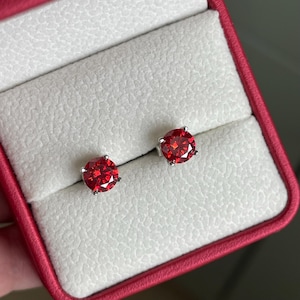May include: A pair of round-cut, vibrant red stud earrings set in silver-toned metal prongs. The earrings are displayed side-by-side, nestled within the white, textured velvet-like interior of a small, bright red jewelry presentation box.