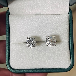 Peut inclure: Une paire de boucles d'oreilles à clous avec des pierres rondes taille brillant serties dans des griffes en métal argenté. Les boucles d'oreilles sont présentées côte à côte dans un écrin vert foncé avec un intérieur blanc texturé.