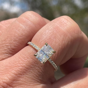 May include: A gold engagement ring with a large, rectangular-cut diamond. The band is embellished with smaller diamonds, enhancing the sparkle. The main diamond is held in place by four prongs. The ring is a classic design.
