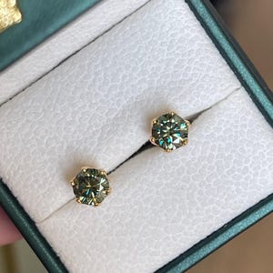May include: A pair of round-cut stud earrings featuring dark, iridescent, greenish-grey stones set in gold-toned metal prongs. The earrings are displayed side-by-side, secured within the white, textured interior lining of a dark green jewellery box.