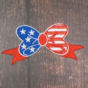 May include: A red, white, and blue bow with a ribbon. The bow is made of fabric and has a star pattern on one side and stripes on the other.