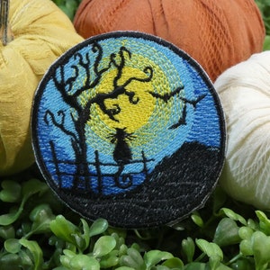 May include: Embroidered iron-on patch featuring a black cat silhouette sitting on a fence under a yellow moon and blue sky. The patch is surrounded by three pumpkins.