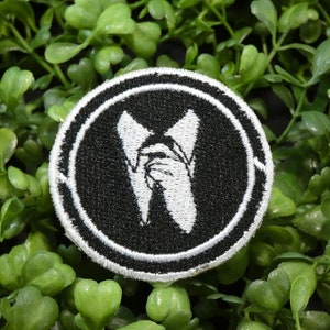May include: A round, black and white embroidered patch featuring a stylized image of a hand holding a pair of scissors. The patch is surrounded by a white border and set against a background of green foliage.