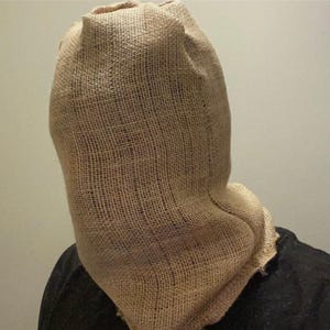 Puede incluir: Una máscara de saco de yute, que cubre la cabeza y el cuello. La máscara es de color marrón claro, con una textura gruesa y tejida. La persona lleva una camisa negra.