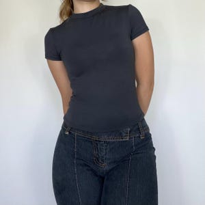 May include: A dark gray, short-sleeved t-shirt with a crew neck. The shirt is paired with dark wash denim jeans. The jeans have a unique design with visible seams and a low-rise waist. The outfit is simple and casual.