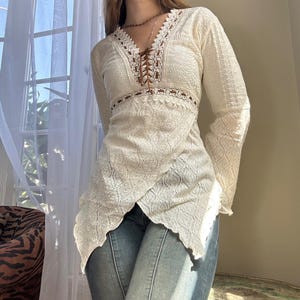 May include: Cream-coloured long-sleeved top with a V-neck and intricate lace detailing. The top features a textured fabric and a unique asymmetrical hemline. The neckline has a brown lace-up design. The sleeves are long and flared.