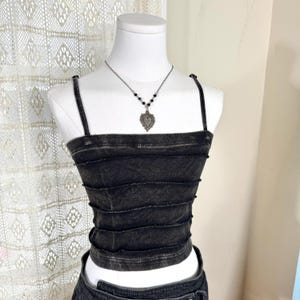 May include: A charcoal gray, acid-washed crop top featuring a square neckline, thin spaghetti straps, and horizontal exposed seam detailing across the front. A silver-toned necklace with a heart-shaped pendant rests on the mannequin's neck.