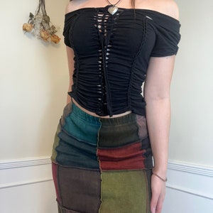 Puede incluir: Top negro con hombros descubiertos con un corpiño fruncido y un detalle de cordones. El top se combina con una falda patchwork en tonos verdes, marrones y rojos. La falda tiene una cintura alta y una silueta ajustada.