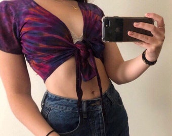May include: A purple and orange tie-dye crop top with a deep V-neck and short sleeves. The top is tied at the front and has a relaxed fit.
