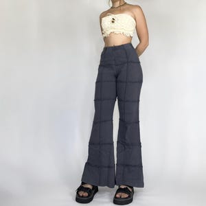 Puede incluir: Pantalones acampanados de color gris oscuro con un diseño de patchwork. Los pantalones tienen una cintura alta y una pernera ancha, con costuras visibles que crean un patrón de rejilla. La persona lleva sandalias de plataforma negras.