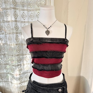 May include: A layered crop top in shades of red and charcoal gray, displayed on a mannequin. The top features a distressed, raw-edge design. A silver necklace with a heart pendant is also visible. The overall aesthetic is edgy and alternative.