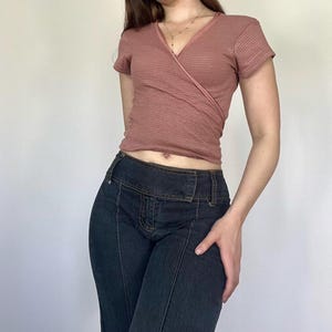 Może przedstawiać: Krótki top z krótkim rękawem w kolorze stonowanego różu z subtelnym wzorem w paski. Top jest połączony z dżinsami z ciemnego denimu. Dżinsy mają wysoki stan i unikalny detal szwu z przodu każdej nogawki.