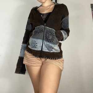 May include: A brown, blue and grey patchwork hoodie with a zip fastening and a hood. The hoodie features various embroidered designs, including eyes, suns and flowers.