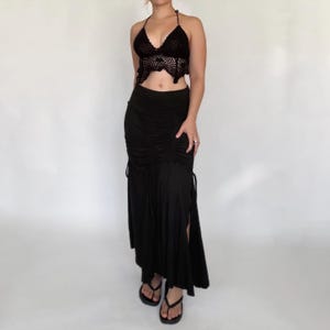 May include: A black crochet halter top with a scalloped hem paired with a matching black maxi skirt featuring a high-waisted design, vertical ruched detailing down the center, and side drawstring ties that create a gathered effect along the legs.