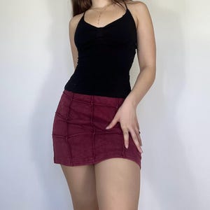 Puede incluir: Un top negro con cuello halter combinado con una minifalda burdeos. La falda presenta un diseño de patchwork con costuras visibles. El conjunto es informal y adecuado para el clima cálido. El top tiene un escote en V.