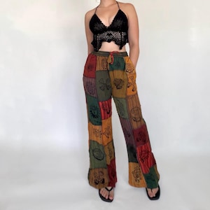 May include: A black crochet halter top with a pointed hem paired with loose-fitting patchwork trousers. The trousers feature a multi-coloured design in shades of green, red, and yellow, decorated with black sun and floral patterns throughout.