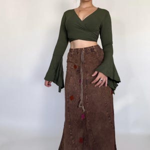 May include: An olive green long-sleeve wrap top with flared sleeves paired with a brown acid-wash maxi skirt. The skirt features a drawstring waist, vertical paneling, and small embroidered diamond-shaped accents in red and purple hues.