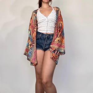 May include: A colorful, patterned kimono-style top with long sleeves. The top features a vibrant mix of red, orange, blue, and gold hues. Worn over a white halter top and denim shorts, it creates a bohemian aesthetic.