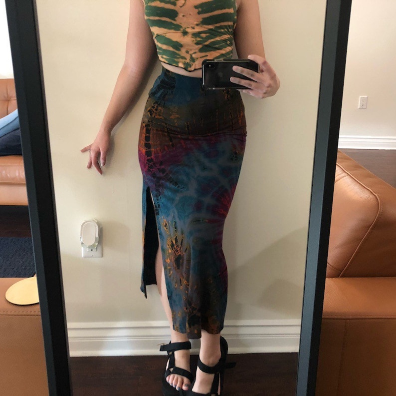 May include: A tie-dye maxi skirt with a high slit, featuring a blend of blue, purple, and brown hues. The skirt is paired with a green and orange tie-dye crop top. Black sandals complete the outfit, reflected in a mirror.