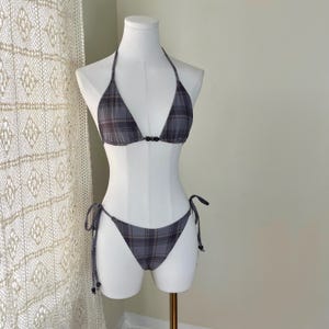 May include: A grey plaid bikini set on a mannequin. The halterneck top has a triangle shape with a small black clasp. The bottoms are a classic bikini cut with tie sides. The fabric has a grey, black, and brown plaid pattern.