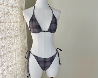 May include: A grey plaid bikini set on a mannequin. The halterneck top has a triangle shape with a small black clasp. The bottoms are a classic bikini cut with tie sides. The fabric has a grey, black, and brown plaid pattern.