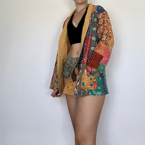 Puede incluir: Una chaqueta de patchwork colorida con un patrón amarillo, rojo, verde y azul. La chaqueta está abierta y tiene un estilo largo y fluido. La persona que lleva la chaqueta también lleva un top corto negro y pantalones cortos de mezclilla.