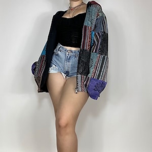 Puede incluir: Una chaqueta de patchwork multicolor con cierre de cremallera. La chaqueta está hecha de diferentes telas y tiene una variedad de patrones, incluyendo rayas, diseños geométricos y motivos florales. La chaqueta se lleva sobre un top negro sin mangas y pantalones cortos de mezclilla.