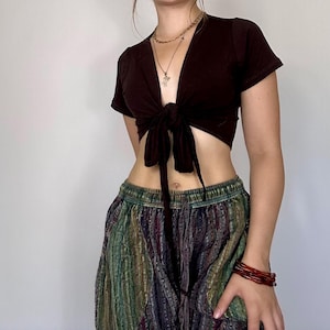 Puede incluir: Un top corto marrón oscuro con lazo y mangas cortas. El top se combina con pantalones sueltos estampados en tonos verdes, azules y morados. Una pila de pulseras rojas adorna la muñeca.
