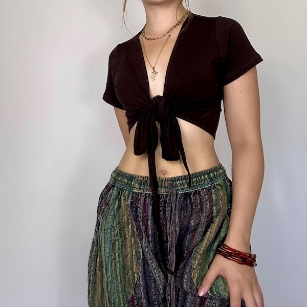 Könnte beinhalten: Ein dunkelbraunes Crop-Top zum Binden mit kurzen Ärmeln. Das Oberteil wird mit einer gemusterten, locker sitzenden Hose in Grün-, Blau- und Lilatönen kombiniert. Ein Stapel roter Armbänder schmückt das Handgelenk.