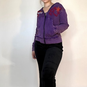 May include: A purple hooded sweatshirt with embroidered floral designs. The sweatshirt is paired with black pants.
