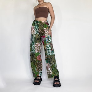 May include: Wide-leg patchwork trousers in green, featuring floral and paisley designs. The trousers incorporate a mix of colours, including green, white, and red. The model is wearing black sandals.