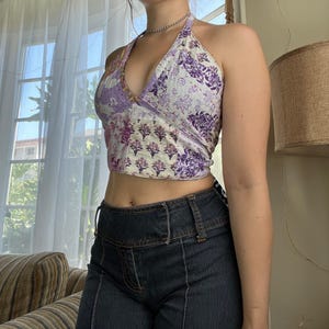 Könnte beinhalten: Ein Neckholder-Top mit Patchwork-Design in Lila-, Lavendel- und Cremetönen. Das Top hat florale und geometrische Muster. Das Model trägt eine dunkelblaue Jeans.