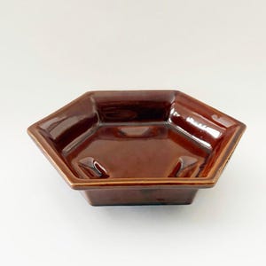 May include: A hexagonal brown ceramic ashtray with a glossy finish. The ashtray has a shallow basin and three curved rests for cigarettes. The edges are slightly raised, and the overall design is simple and functional.