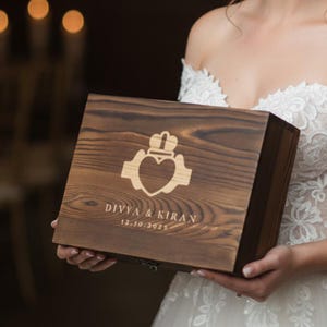 May include: A wooden box with a Claddagh design, heart, crown, and hands, engraved on the lid. The box is a dark brown color and has the names "DIVYA & KIRAN" and the date "12.10.2025" engraved on it. The box is held by a person wearing a white dress.