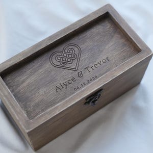 Puede incluir: Una caja rectangular de madera con tapa con bisagras. La caja está teñida de color marrón medio. La tapa está grabada con un diseño de nudo en forma de corazón y los nombres "Alyce & Trevor" y la fecha "04.12.2030".