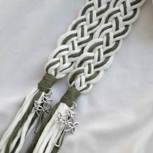 May include: Braided white and olive green macrame cord with silver-colored Celtic knot charms. The cord is tightly woven, with a tassel at the end. The background is a soft, off-white fabric.